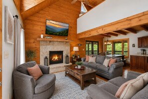 A cozy living room with a stone fireplace, wooden beams, and a large window overlooking greenery. - Mountain Escape - Hot Tub - Game Room - Sleeps 8 (Pigeon Forge)