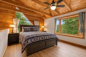 A bedroom with a wooden ceiling, a bed with patterned bedding, a nightstand, a window with curtains, and a view of greenery outside. - Mountain Escape - Hot Tub - Game Room - Sleeps 8 (Pigeon Forge)