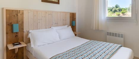 Un dormitorio con cabecera de madera, cama con ropa de cama blanca, manta a rayas, mesita de noche con un libro y ventana con vista a follaje.