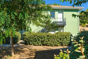 A two-story building with a balcony, surrounded by dense shrubbery and trees.