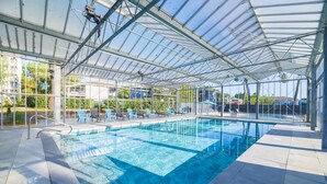 A covered swimming pool with clear blue water, surrounded by lounge chairs and a glass ceiling structure.