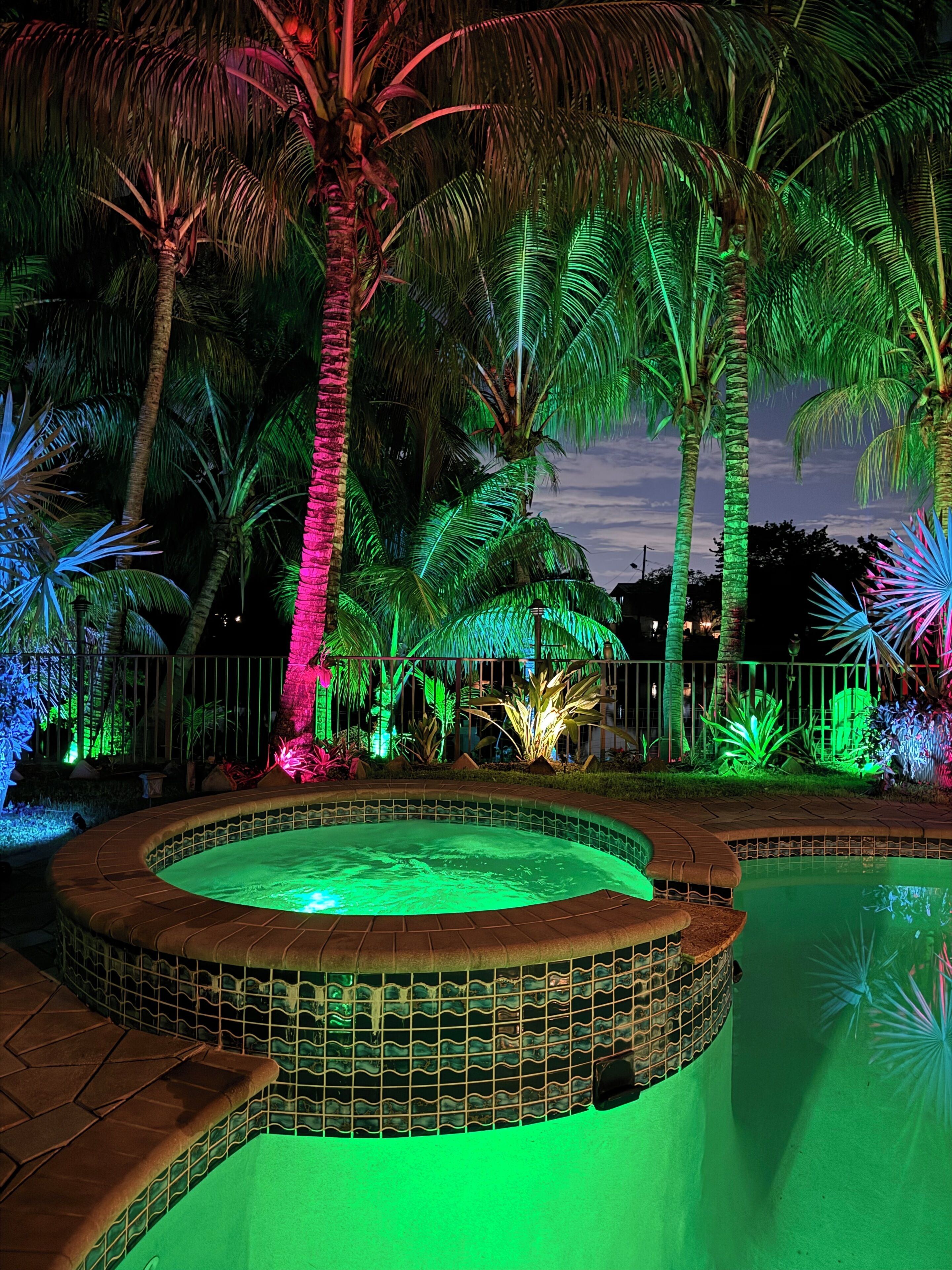 Een terras buiten, 's nachts, met een rond zwembad, palmbomen en decoratieve verlichting.