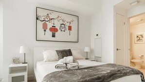 Ein Schlafzimmer mit einem Bett, zwei Nachttischen, einem Wandregal und einem Bild über dem Bett. Eine Tür führt ins Badezimmer.