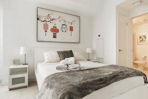 Un dormitorio con cama, dos mesitas de noche, un buró y una obra de arte enmarcada sobre la cama, además de una puerta que da al baño.