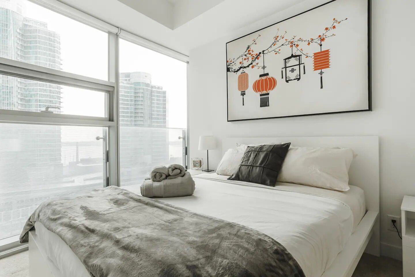 Een moderne hotelkamer met een groot raam, een net opgemaakt bed, een decoratief wandobject en een nachtkastje met een lamp.
