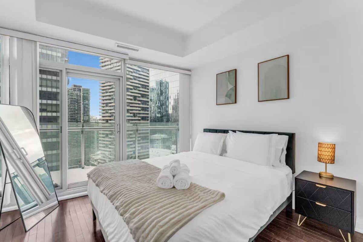 A modern hotel room with a large bed, a bedside table with a lamp, and a view of the city through floor-to-ceiling windows.