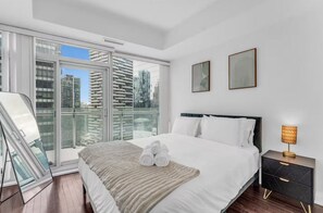 Quarto de hotel moderno com uma cama grande, uma mesinha de cabeceira com abajur e vista da cidade através de janelas do chão ao teto.