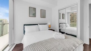 A modern hotel room with a large bed, a bedside table, a nightstand, a window with a view of buildings, and a door leading to another room. - Harbour Skyline (Toronto)