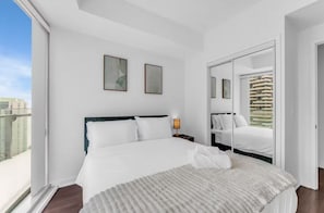 Una habitación de hotel moderna con una cama grande, una mesita de noche, una ventana con vistas a edificios y una puerta que da a otra habitación.