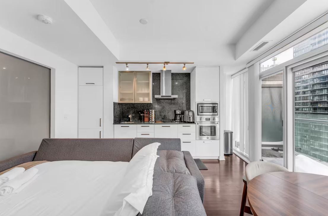 A modern hotel room with a large bed, a sofa, a dining table, and a kitchenette.