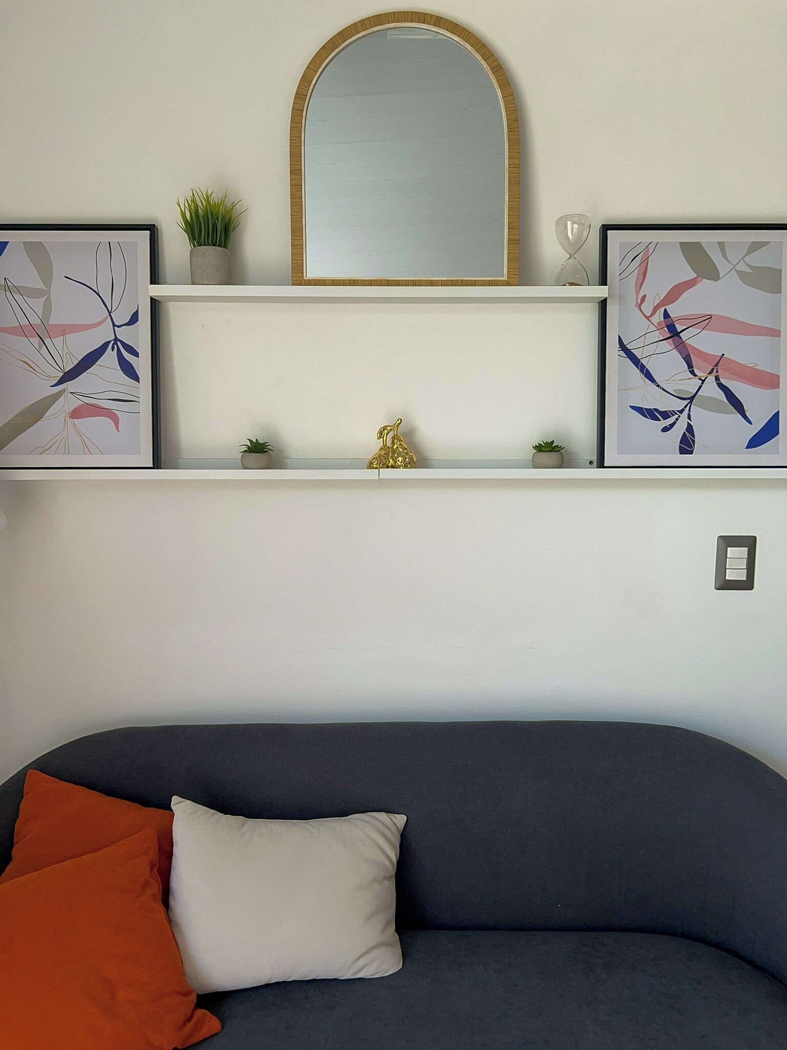 A modern living room with a dark grey sofa, a white cushion, and a red cushion. Above the sofa, there are two framed abstract paintings and a small potted plant on a white shelf. A gold decorative item is placed on the shelf.