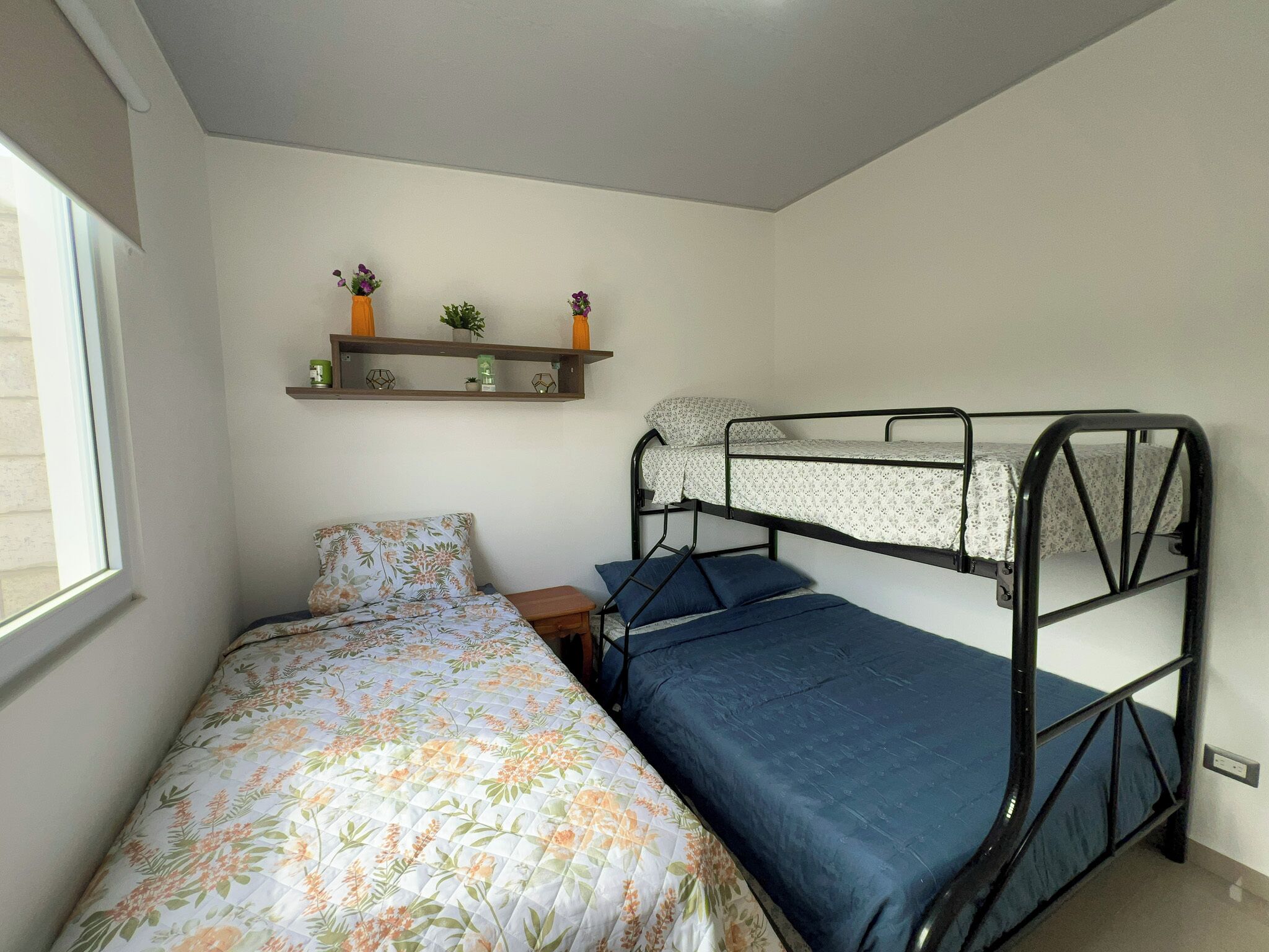 Ein Schlafsaalzimmer mit Etagenbett, einem Einzelbett mit floralem Bettbezug, einem kleinen Nachttisch aus Holz und einem Regal mit dekorativen Gegenständen.