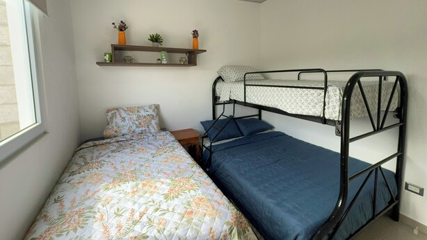 Un dormitorio con una cama litera, una cama individual con ropa de cama floral, una pequeña mesita de noche de madera y una estantería con adornos.