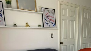 Un salón con un sofá azul oscuro, sillones naranjas, una mesa de centro blanca y una mesita auxiliar blanca. Se observan cuadros enmarcados en la pared, una planta en maceta y una puerta al fondo.
