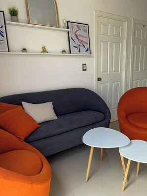 Ein Wohnzimmer mit einem dunkelblauen Sofa, orangefarbenen Sessel und einem weißen Couchtisch sowie einem kleinen weißen Tisch. An der Wand befinden sich gerahmte Bilder, eine Zimmerpflanze und im Hintergrund eine Tür.