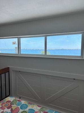 A room with a view of a body of water through a window, a white radiator, and a colorful quilt on the bed.