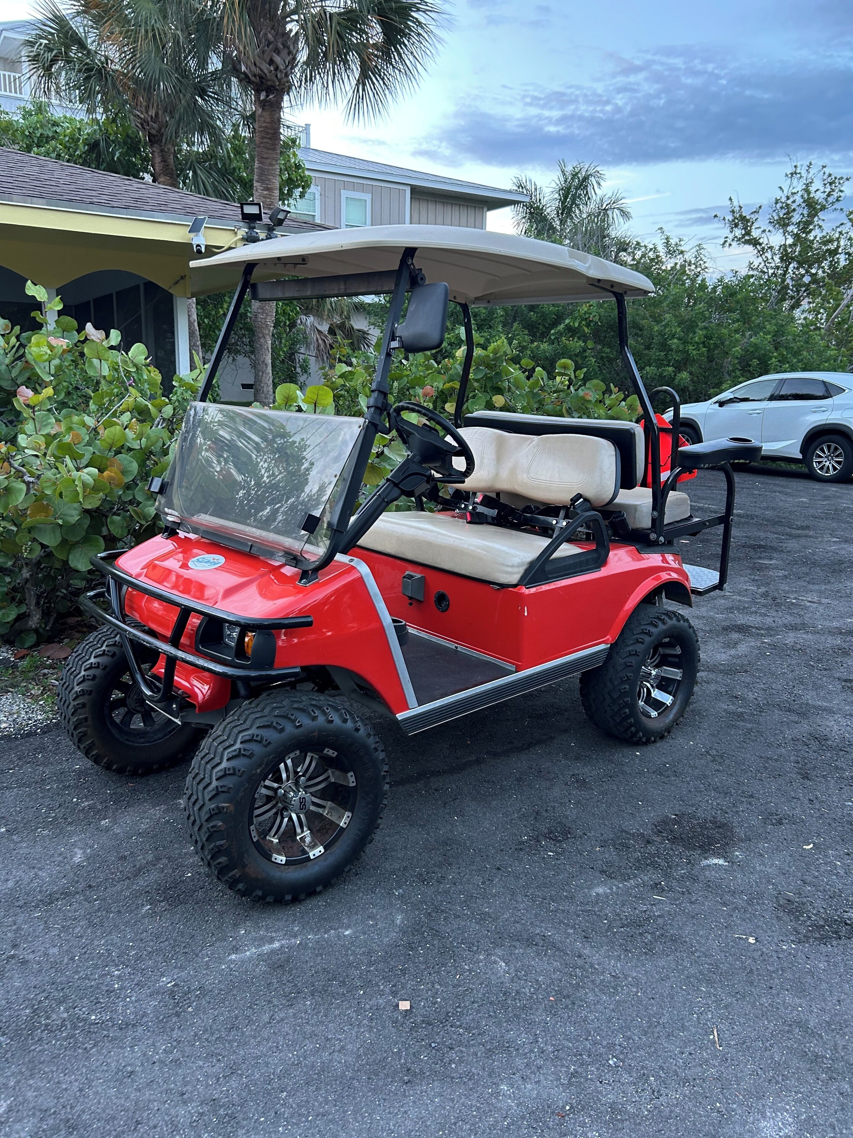 Une voiturette de golf rouge est garée sur une surface pavée, avec une maison et des palmiers en arrière-plan.