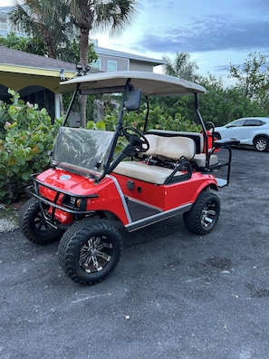 A red golf cart parked on a paved surface with a house and palm trees in the background.