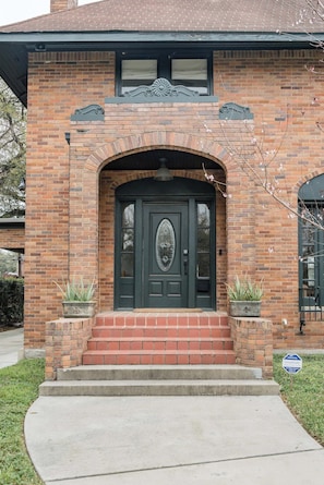 A two-story brick house with a dark green front door, a small porch with steps, and two potted plants on either side. - Historic Houston Midtown Haven (Houston)