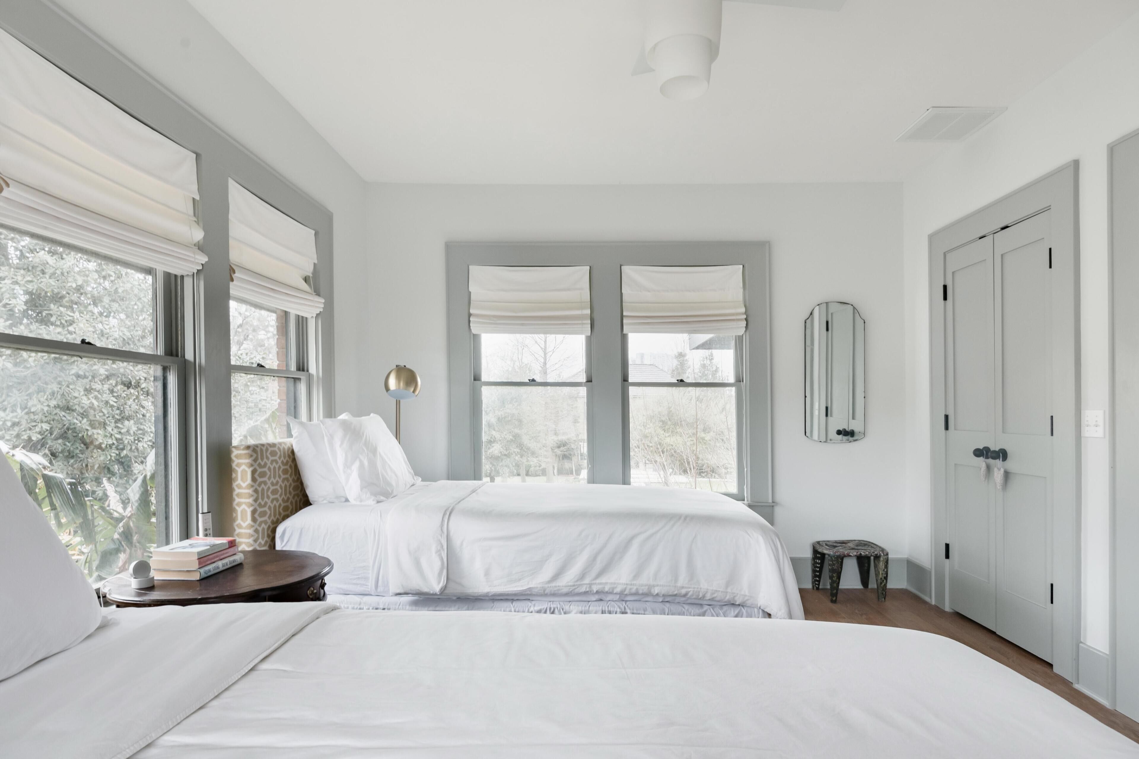 A bedroom with a large bed, two windows with white blinds, a bedside table with books, and a small stool.