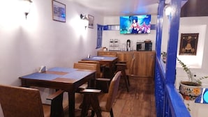 Un comedor con mesas y sillas de madera, un televisor montado en la pared y una cocina con microondas y cafetera.