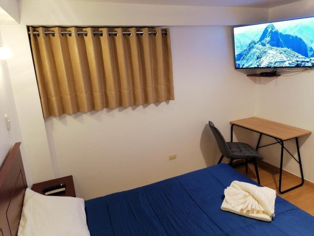 Habitación de hotel con cama, escritorio, silla, televisor y cortina.