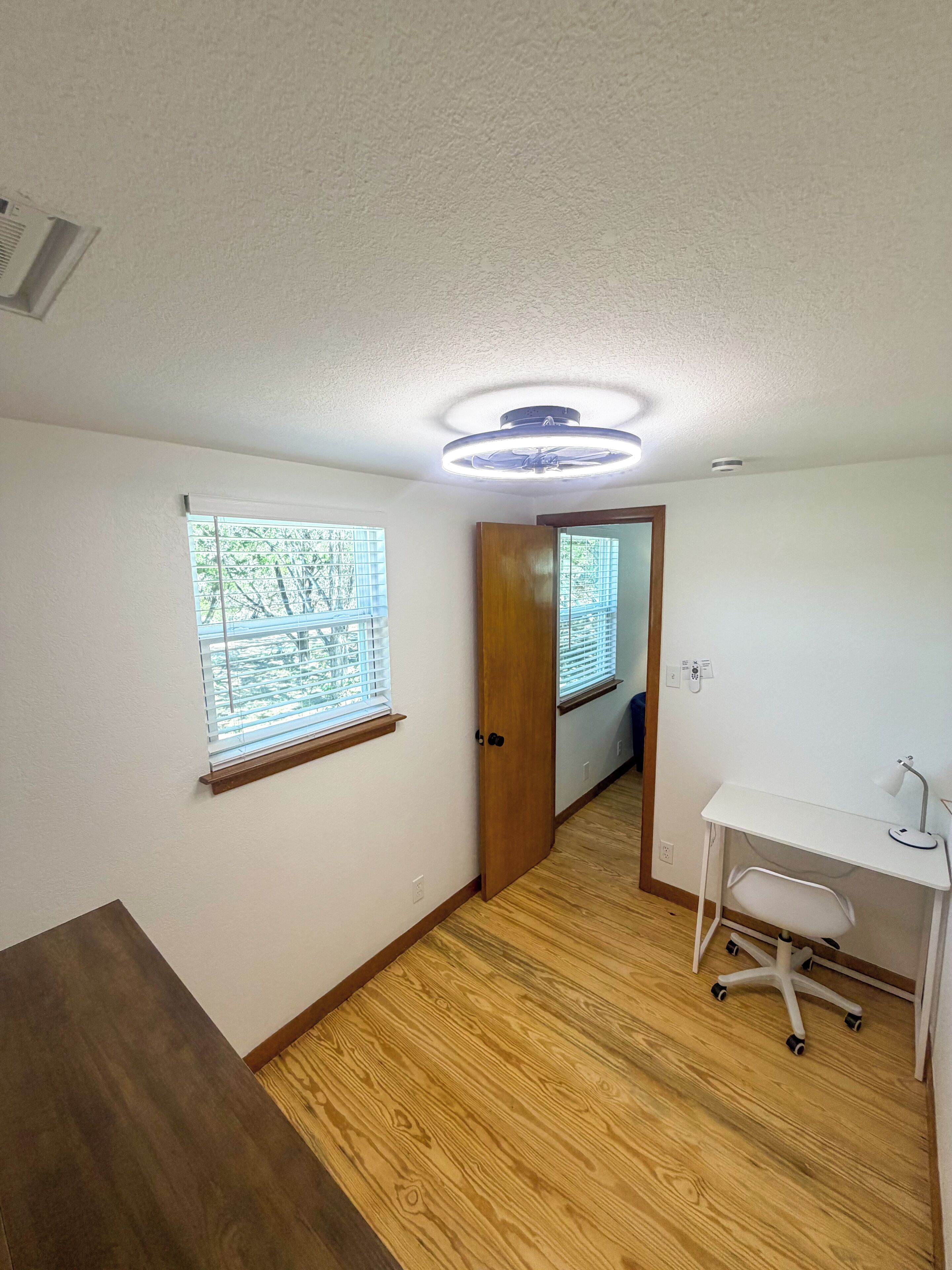 A room with a wooden floor, a white desk with a chair, and two windows with blinds.