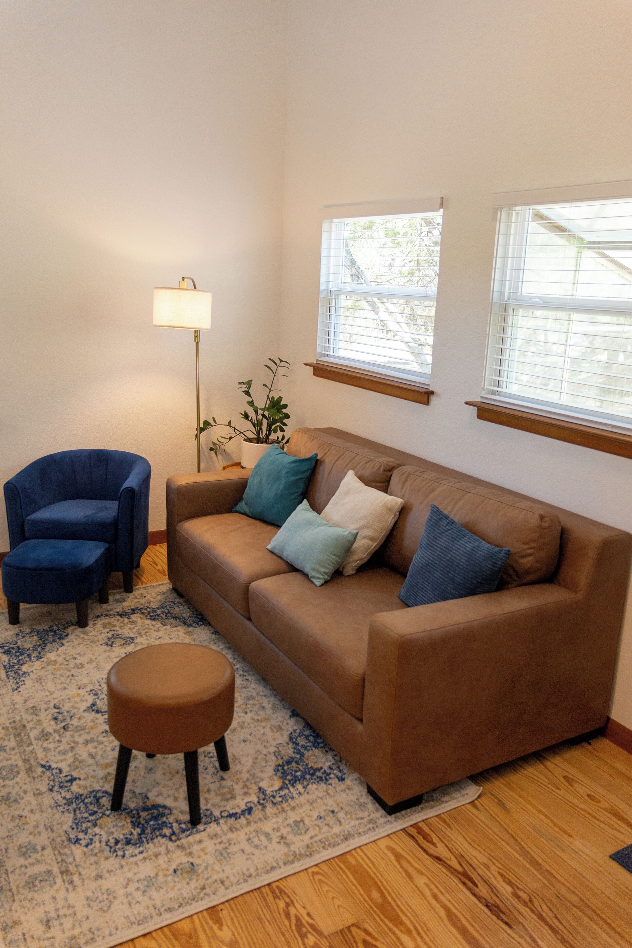 A living room with a brown sofa, two blue armchairs, a small round ottoman, a floor lamp, and a potted plant.