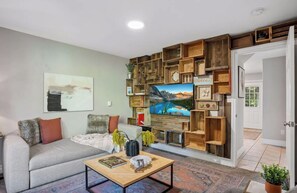 Ein Wohnzimmer mit einer Couch, einem Couchtisch und einem an der Wand befestigten Fernseher. Eine Tür führt in einen anderen Raum.