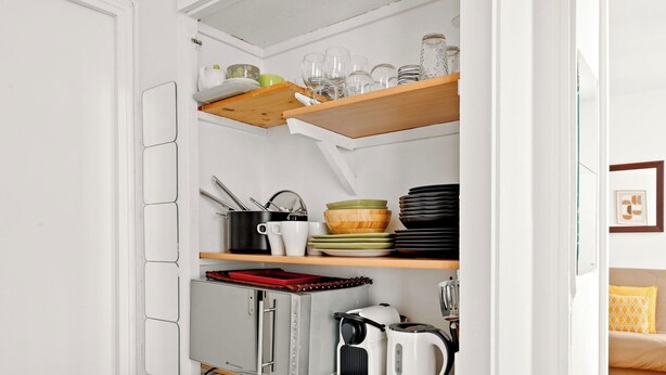 Una cocina con armarios blancos, una encimera con hervidor, cafetera y una bandeja roja. Hay estantes con cristalería y platos.