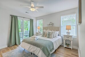 A bedroom with a bed, a ceiling fan, a nightstand, a lamp, and a view of the outdoors through a window. - Pilgrim Luxury #4. New Construction with a Hot tub (Hanahan)