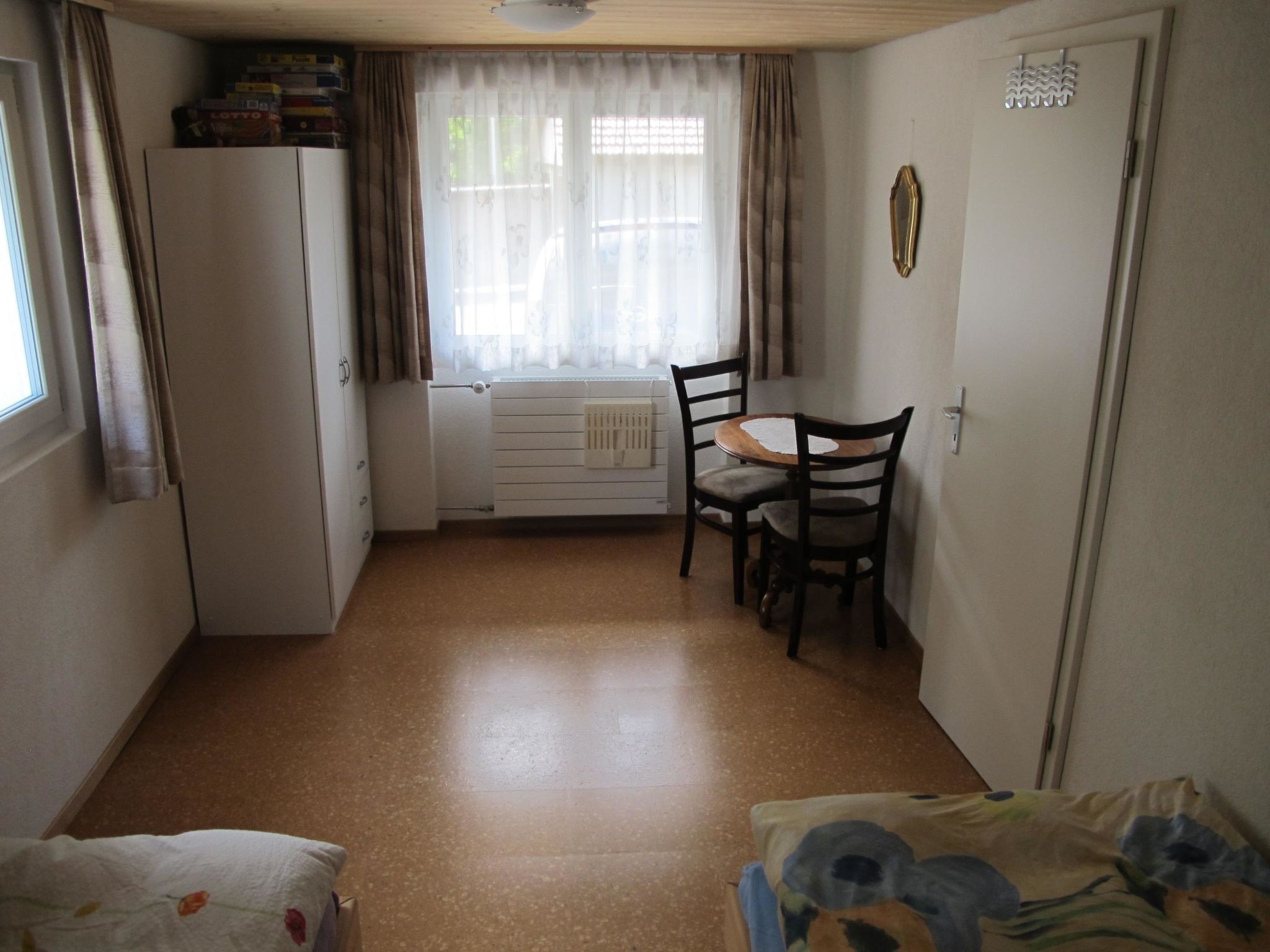Una habitación pequeña con cama, una mesa y sillas, un radiador y un ventanal con cortinas.