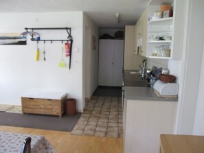 Eine kleine Küche mit Spüle, Schränken und einer Arbeitsplatte. Ein Holzbänkchen und ein Mülleimer stehen im Raum.