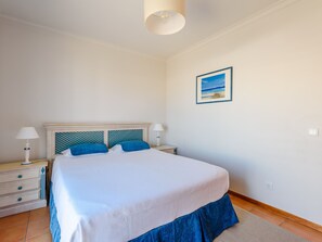 Quarto com cama, duas mesas de cabeceira, um criado-mudo, abajur e um quadro na parede.