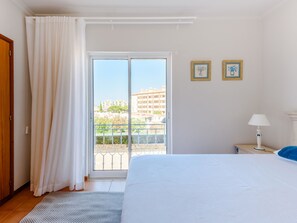 Ein Hotelzimmer mit einem Bett, einem Nachttisch, einer Lampe, einem Fenster mit Vorhängen und zwei gerahmten Bildern an der Wand.