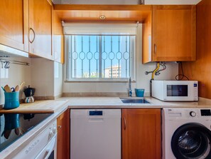 Cozinha compacta com armários de madeira, máquina de lavar louça embutida, máquina de lavar roupa, micro-ondas e janela com grade de metal.