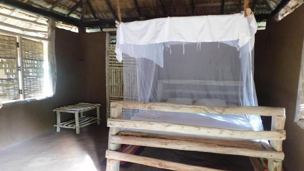 Un simple lit en bois, surmonté d’une moustiquaire, dans une pièce dont le toit est fait de chaume et les murs en bois.
