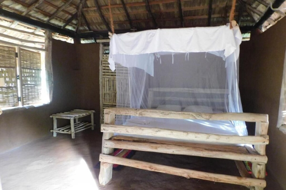A simple wooden bed with a mosquito net in a room with a thatched roof and wooden walls.