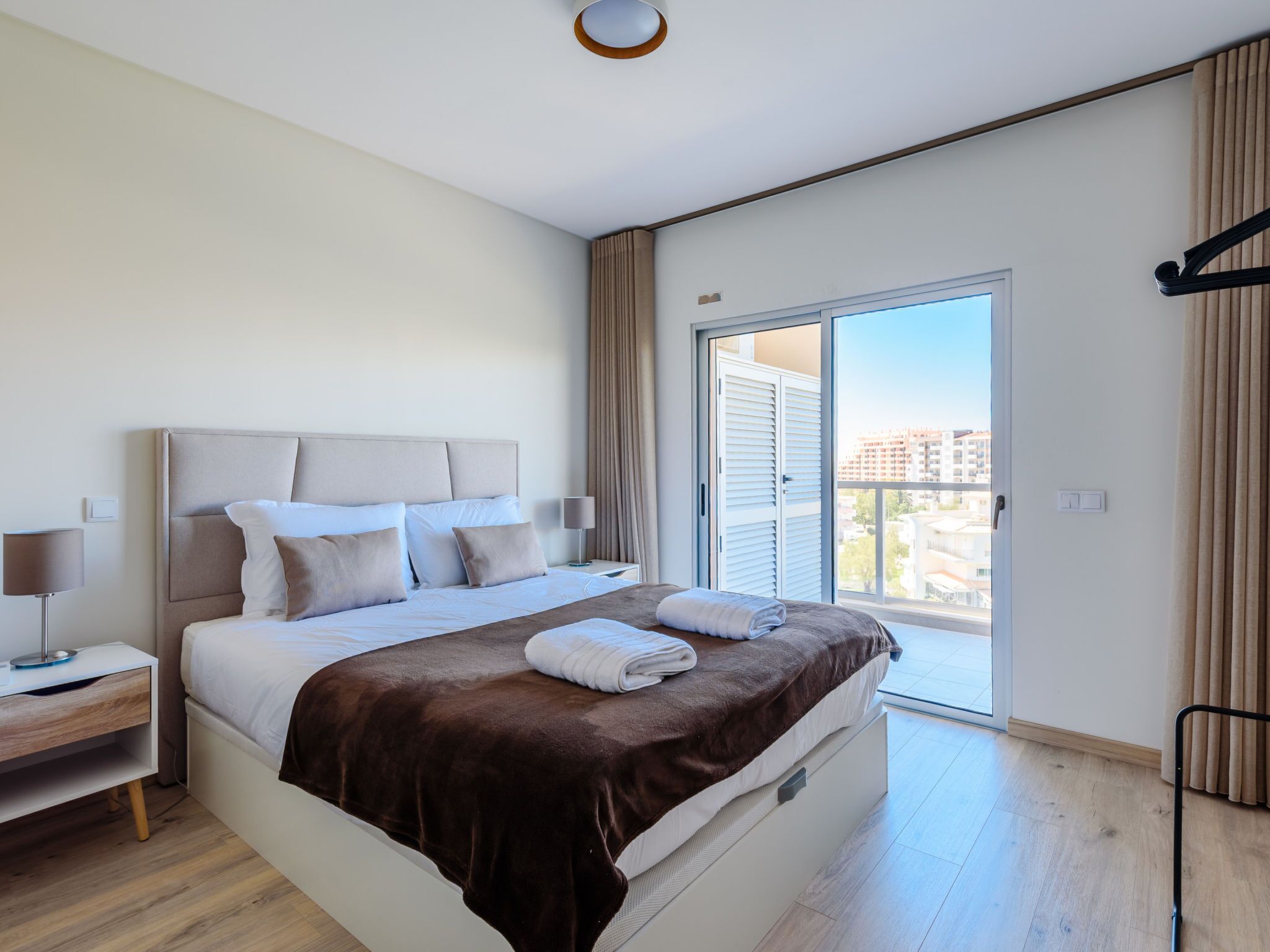 Quarto de hotel moderno com uma cama grande, uma mesinha de cabeceira, uma luminária e vista para a cidade por uma porta de correr.