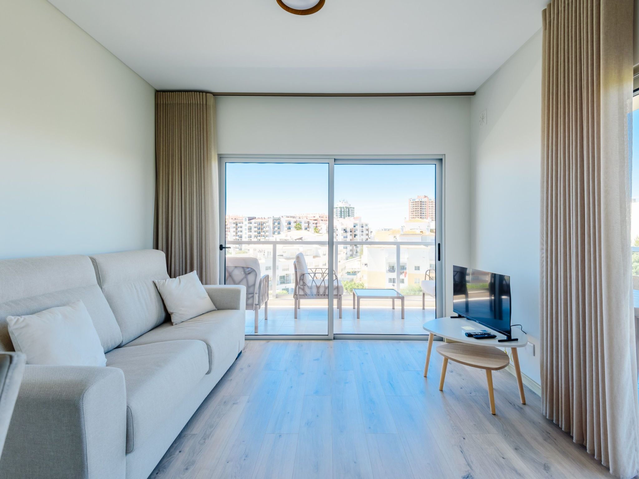 A modern living room with a sofa, a TV on a stand, and a balcony with a view of buildings.