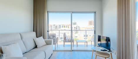 A modern living room with a sofa, a TV on a stand, and a balcony with a view of buildings.