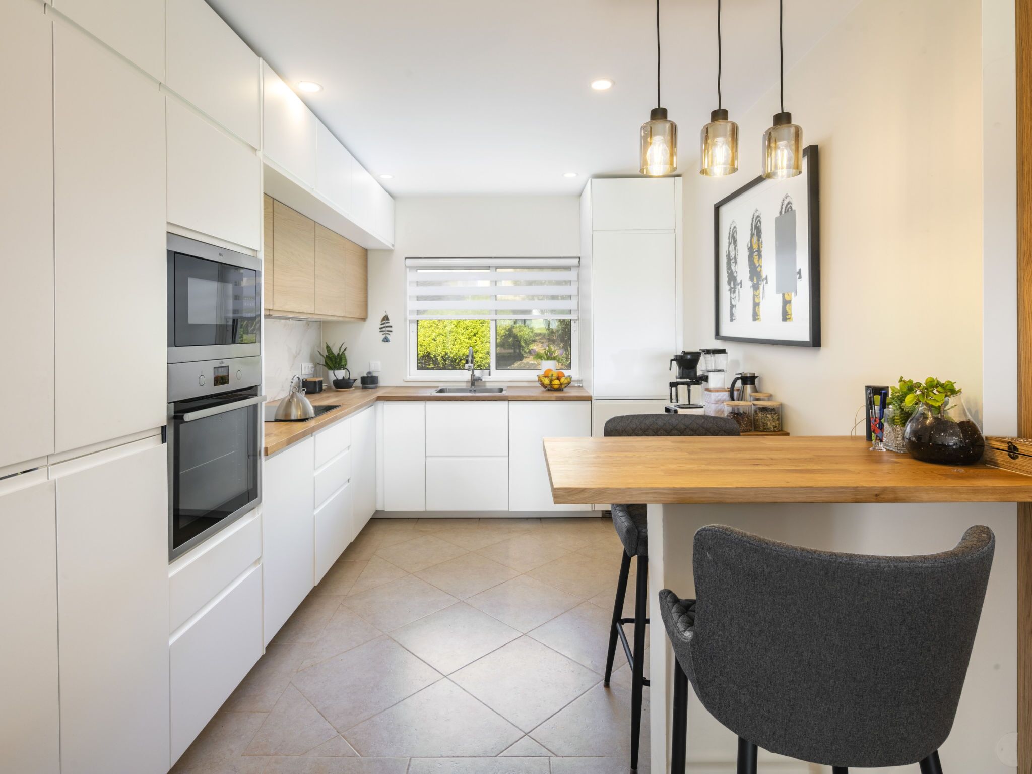 A modern kitchen with white cabinetry, a wooden countertop, and a built-in oven. There is a window with a view of greenery outside, and a bar area with stools.