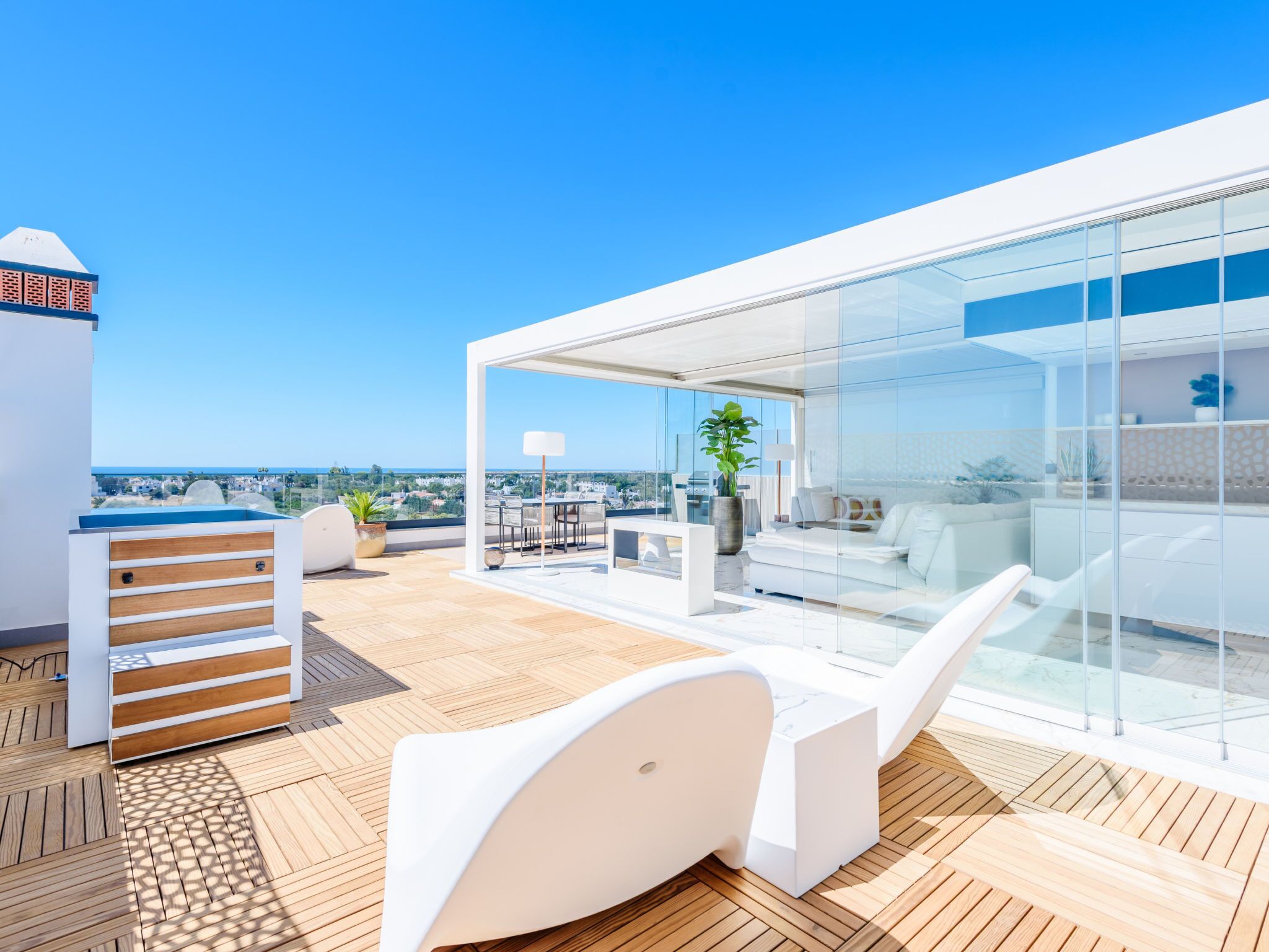 Una terraza moderna en la azotea con cerramiento de cristal, suelo de madera y mobiliario exterior.
