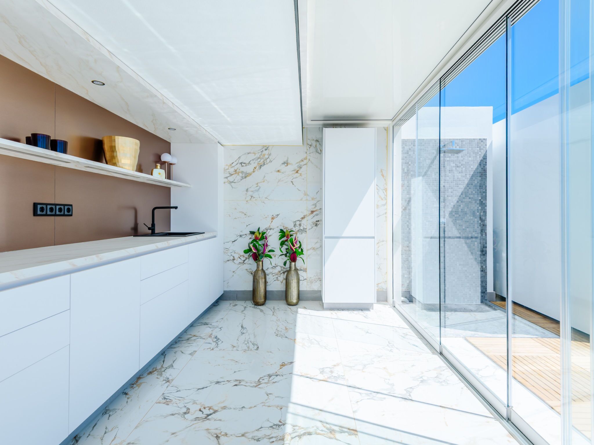 Une cuisine moderne avec un sol en marbre, des meubles blancs et un balcon fermé par des baies vitrées.