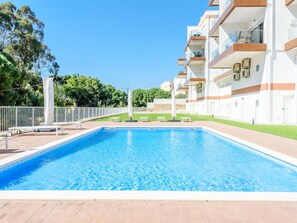 Ein rechteckiger Pool mit Holzdeck im Vordergrund, umgeben von einem weißen Zaun. Im Hintergrund ein mehrstöckiges Gebäude mit Balkonen und einem strahlend blauen Himmel.