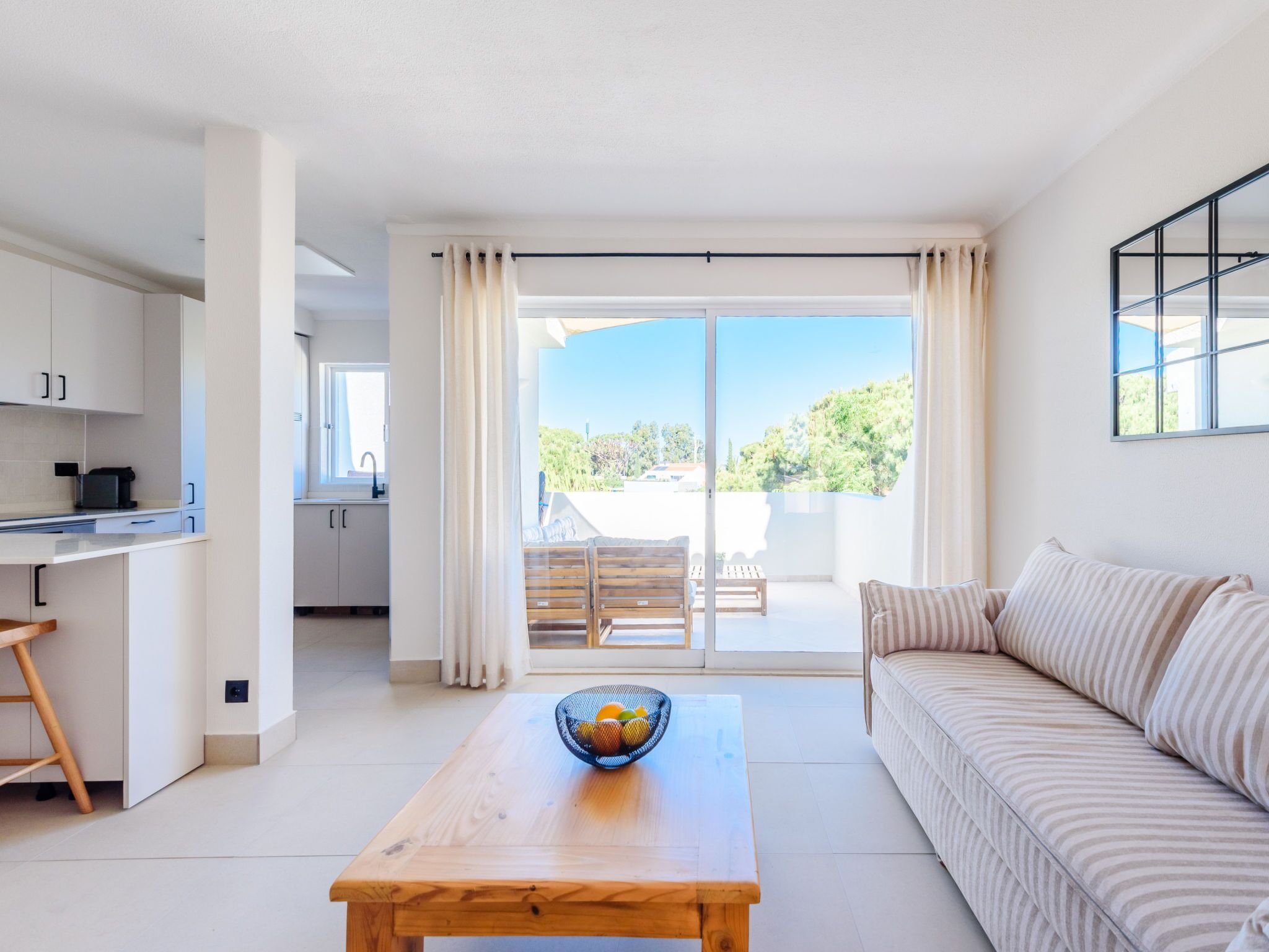 A modern living room with a sofa, a wooden table with a fruit bowl, and a kitchen area with cabinets and a countertop.