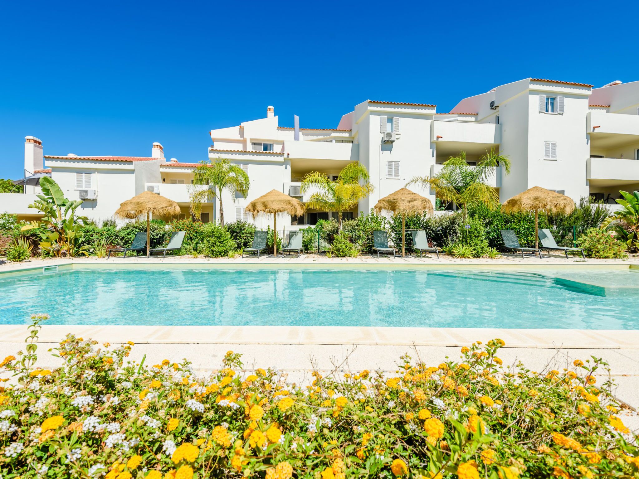 A resort with a swimming pool, white buildings, and thatched umbrellas.