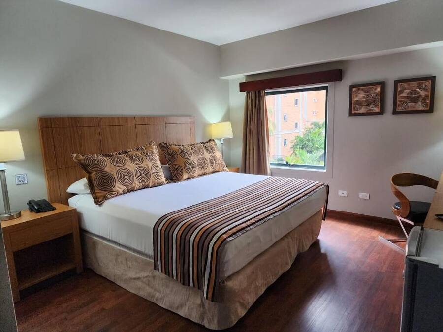 A hotel room with a large bed, a wooden headboard, two bedside lamps, a desk with a chair, and a window with a view of buildings and greenery.