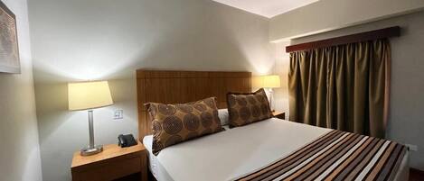 Une chambre d’hôtel avec un lit, deux tables de chevet avec des lampes, une tête de lit et une fenêtre avec des rideaux.