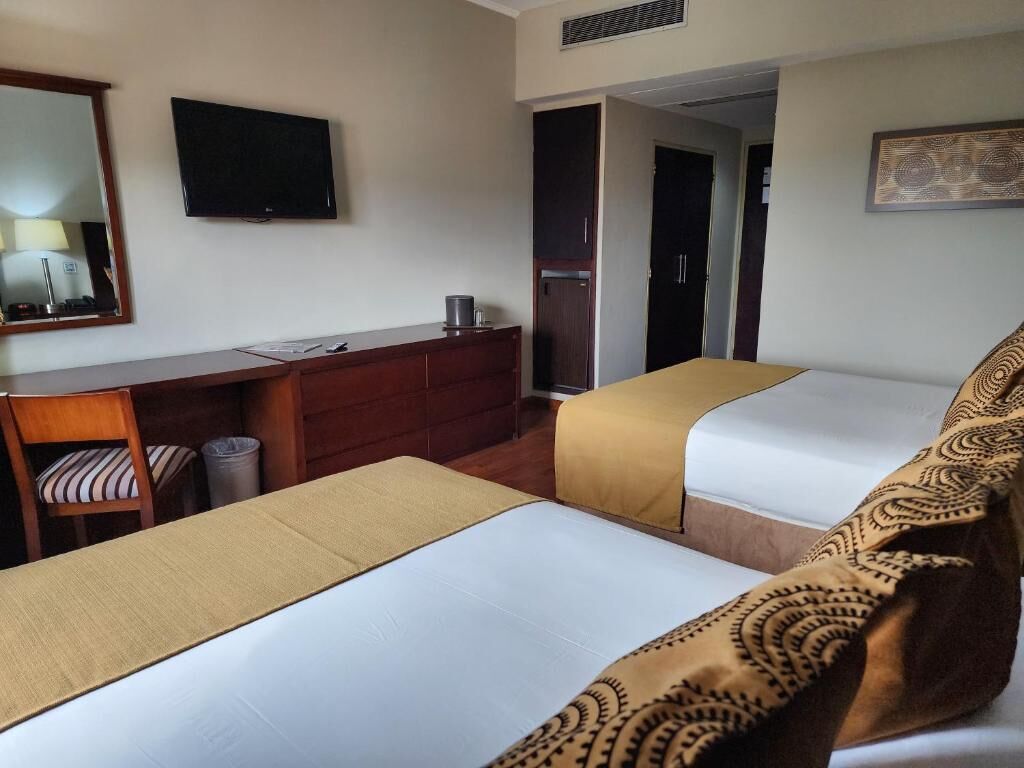 Une chambre d’hôtel avec deux lits, un bureau, une chaise, une télévision et une porte donnant sur une autre pièce.